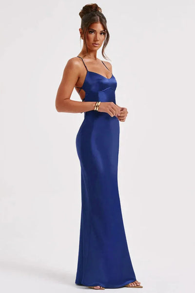A model wearing a sleeveless, V-neck, backless, blue maxi dress with an empire waist, standing in a room with a floral arrangement and a pink column in the background.